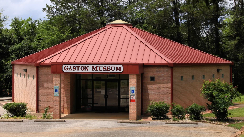 Brick building with a red standing-seam metal roof and a sign that reads 'Gaston Museum' above the entrance, surrounded by green trees.